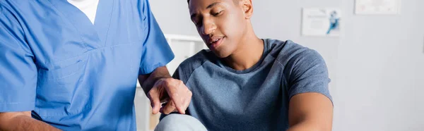 Chiropratico maturo che punta con il dito al ginocchio ferito del paziente afroamericano, striscione — Foto stock