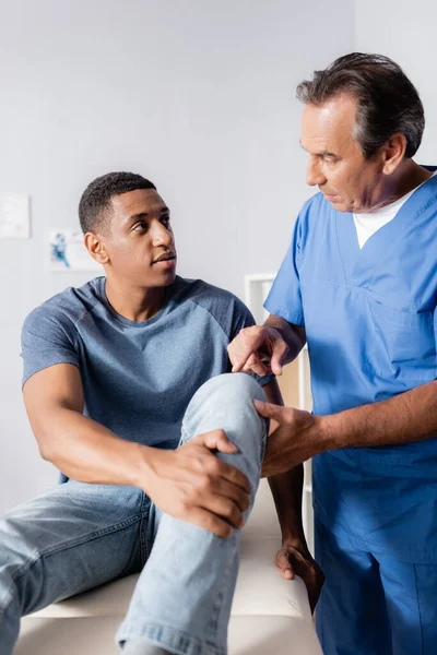 Terapeuta maduro señalando con el dedo a la rodilla lesionada de hombre afroamericano en la clínica - foto de stock