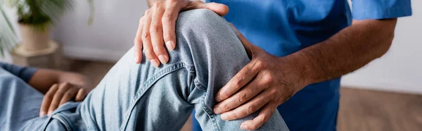 Ausgeschnittene Ansicht eines erfahrenen Therapeuten, der mit dem verletzten Knie eines Afroamerikaners in der Klinik arbeitet, Banner — Stockfoto