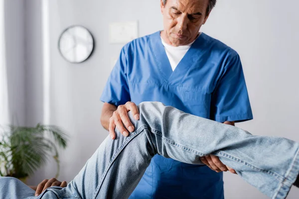 Terapeuta maduro trabajando con rodilla lesionada de hombre afroamericano en clínica - foto de stock