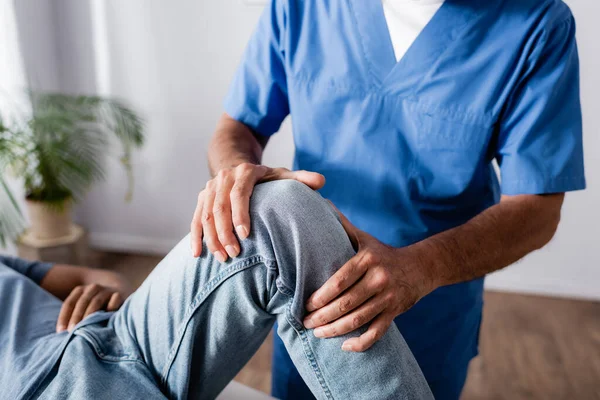 Vista recortada de terapeuta trabajando con rodilla lesionada de hombre afroamericano en clínica - foto de stock