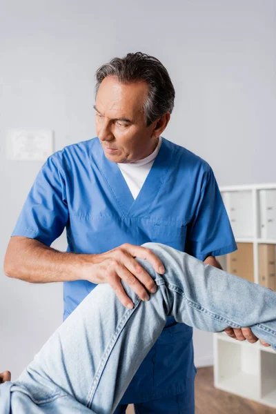 Massaggiatore maturo che lavora con il ginocchio ferito del paziente in clinica — Foto stock