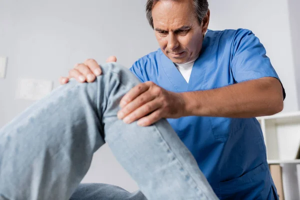 Массажист среднего возраста, работающий с поврежденным коленом пациента в клинике — стоковое фото