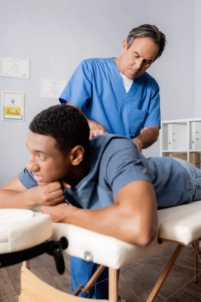 Middle aged chiropractor working with young african american patient on blurred foreground — Stock Photo
