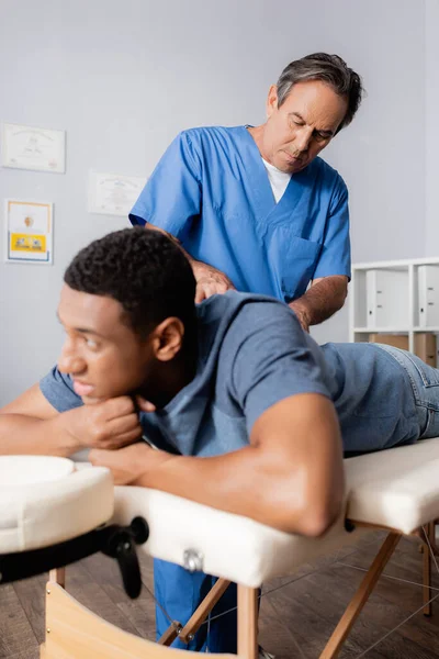 Chiropratico di mezza età che lavora con un giovane paziente afroamericano in primo piano sfocato — Foto stock