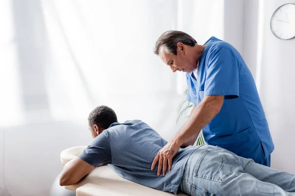 Quiropráctico de mediana edad que trabaja con un paciente afroamericano joven y lesionado en la mesa de masaje en la clínica - foto de stock