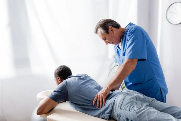 Chiropraktiker mittleren Alters arbeitet mit jungen und verletzten afrikanisch-amerikanischen Patienten auf Massagetisch in Klinik — Stockfoto