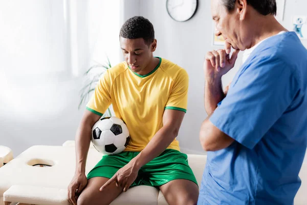 Jugador de fútbol afroamericano lesionado señalando con el dedo en la rodilla cerca del médico en la clínica - foto de stock