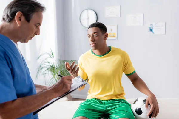 Jugador de fútbol afroamericano lesionado sentado en la mesa de masaje con pelota y gesto cerca del médico en la clínica - foto de stock