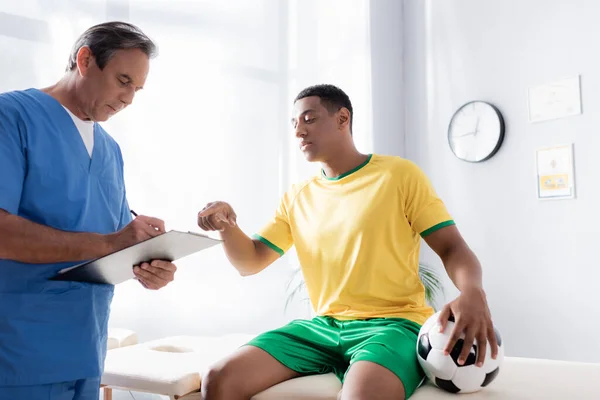 Arzt schreibt Rezept in der Nähe verletzter afrikanischer Fußballspieler zeigt mit Finger in Klinik — Stockfoto