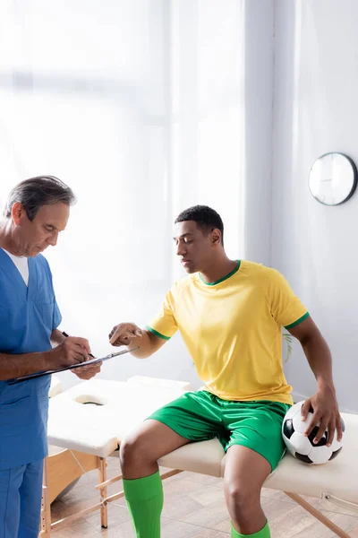 Doctor writing prescription near injured african american football player sitting with ball on massage table — Stock Photo
