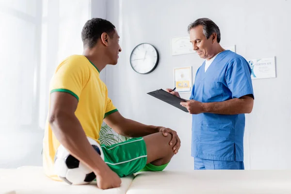 Doctor writing prescription near smiling african american football player sitting with ball on massage table — Stock Photo