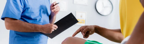Vista parcial del deportista afroamericano lesionado apuntando con el dedo a la rodilla cerca del médico en la clínica, pancarta - foto de stock