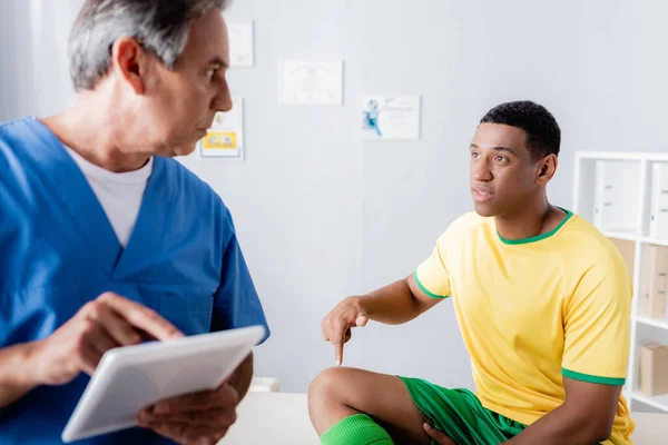 Doctor holding digital tablet and looking at injured african american football player pointing with finger at knee — Stock Photo