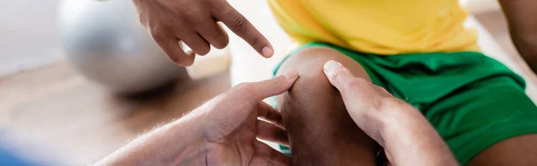 Cropped view of injured african american sportsman pointing with finger at knee near chiropractor, banner — Stock Photo