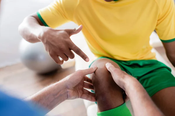 Vista recortada del deportista afroamericano lesionado apuntando con el dedo a la rodilla cerca de quiropráctico - foto de stock