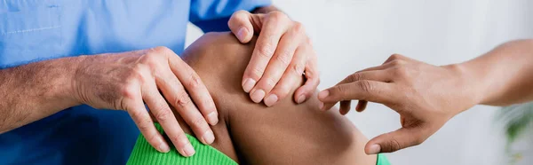 Cropped view of african american sportsman pointing with finger at injured knee near doctor, banner — Stock Photo