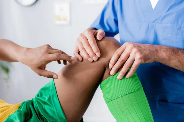 Vista recortada del deportista afroamericano señalando con el dedo a la rodilla lesionada cerca del médico - foto de stock