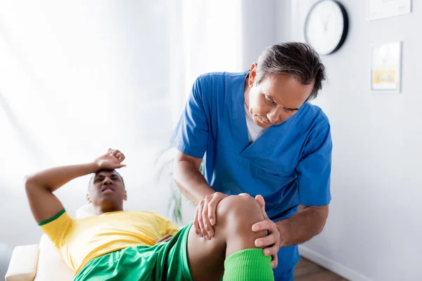 Masseur d'âge moyen massant le genou d'un sportif afro-américain sur fond flou — Photo de stock