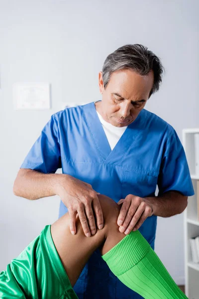 Masseur d'âge moyen massant le genou d'un sportif afro-américain en clinique — Photo de stock