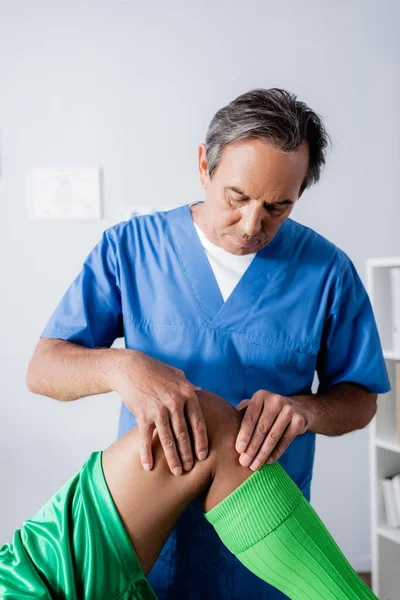 Masajista de mediana edad masajeando rodilla de deportista afroamericano en clínica - foto de stock