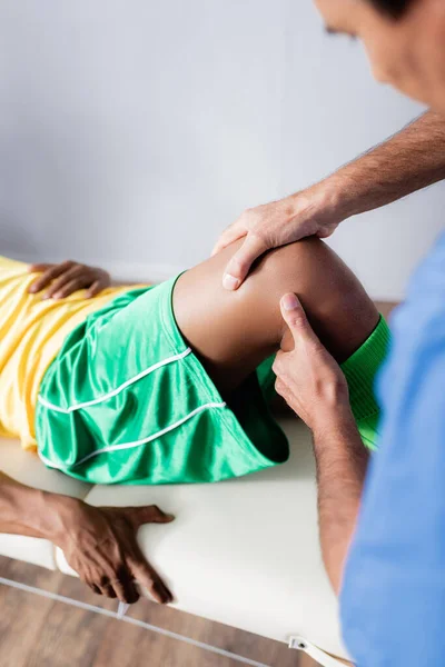 Massothérapeute d'âge moyen flou massant le genou d'un sportif afro-américain en tenue de sport en clinique — Photo de stock