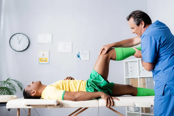 Side view of middle aged masseur working with injured african american sportsman in sportswear in clinic — Stock Photo