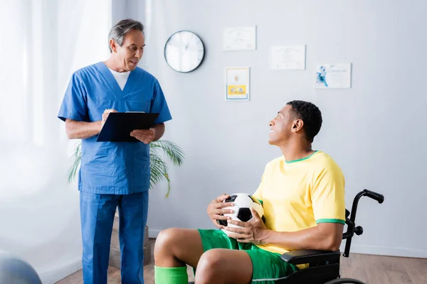 Disabled african american sportsman in wheelchair holding football and smiling near doctor — Stock Photo