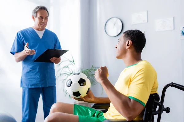 Cheerful african american sportsman in wheelchair holding football near doctor on blurred background — Stock Photo