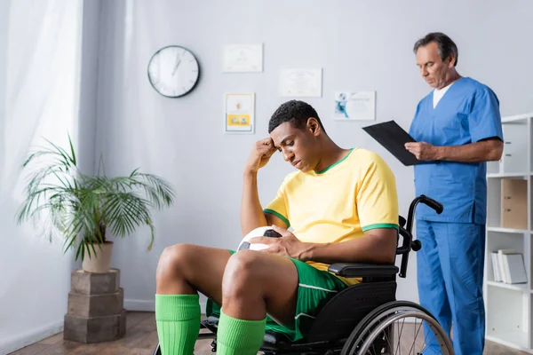 Triste afro-americano desportista em cadeira de rodas segurando futebol perto médico em fundo turvo — Fotografia de Stock