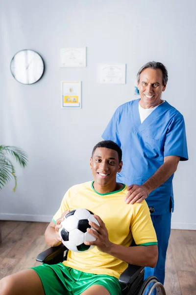 Disabled african american sportsman in wheelchair holding football and smiling with doctor — Stock Photo