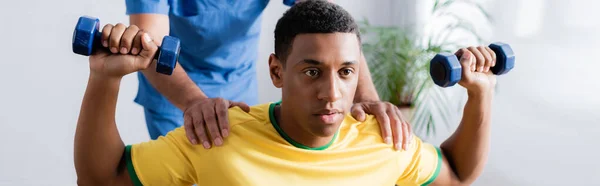 Sportif afro-américain faisant de l'exercice avec des haltères près d'un physiothérapeute sur fond flou, bannière — Photo de stock