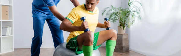 Afrikanisch-amerikanischer Sportler beim Training mit Kurzhanteln auf Fitnessball in der Nähe des Physiotherapeuten, Banner — Stockfoto