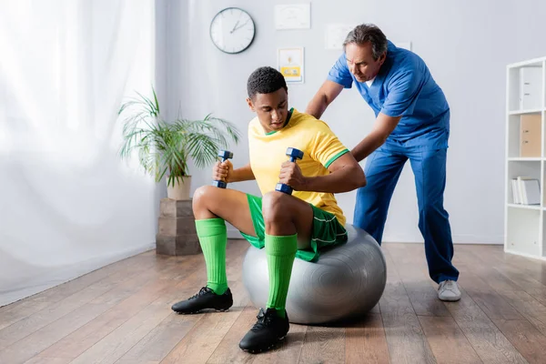 African american sportsman exercising with dumbbells on fitness ball near physiotherapist in clinic — Stock Photo
