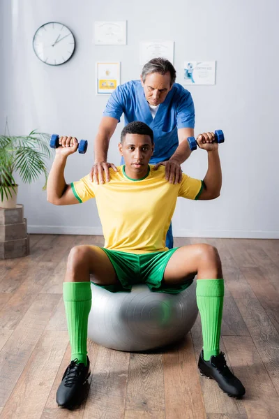 Entrenamiento de deportista afroamericano con pesas en pelota de fitness cerca de fisioterapeuta durante la rehabilitación - foto de stock