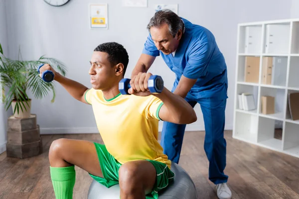 Uomo afroamericano in abbigliamento sportivo formazione con manubri sulla palla fitness vicino fisioterapista — Foto stock