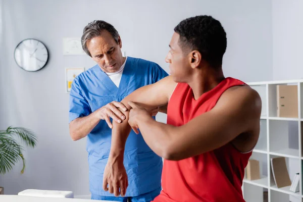 Quiropráctico maduro que trabaja con el hombre afroamericano en ropa deportiva en primer plano borroso — Stock Photo