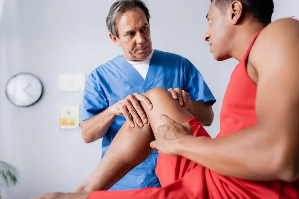 Mature chiropractor working with injured knee of african american man in sportswear on blurred foreground — Stock Photo