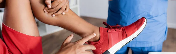 Vista recortada del hombre afroamericano en zapatilla de deporte que apunta con el dedo a la pierna lesionada cerca del médico en la clínica, pancarta - foto de stock