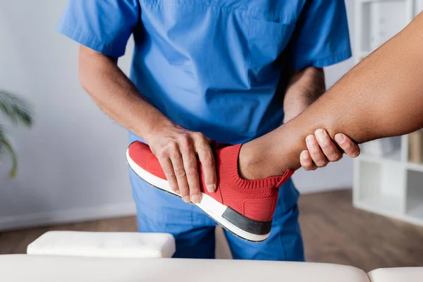 Partial view of chiropractor working with injured keg of african american man in sneaker — Stock Photo