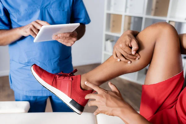 Vista recortada del hombre afroamericano en zapatilla de deporte apuntando con el dedo a la pierna lesionada cerca del médico con tableta digital sobre fondo borroso - foto de stock