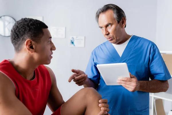 Chiropraktiker hält digitales Tablet während er in der Nähe eines afrikanisch-amerikanischen Patienten in Sportbekleidung spricht und gestikuliert — Stockfoto