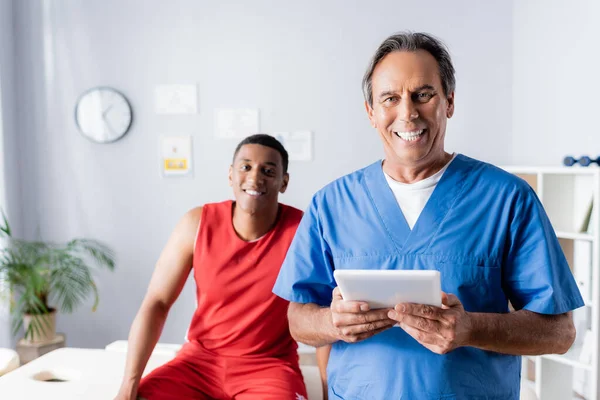 Médico alegre segurando tablet digital perto de paciente americano africano feliz em sportswear em fundo borrado — Fotografia de Stock