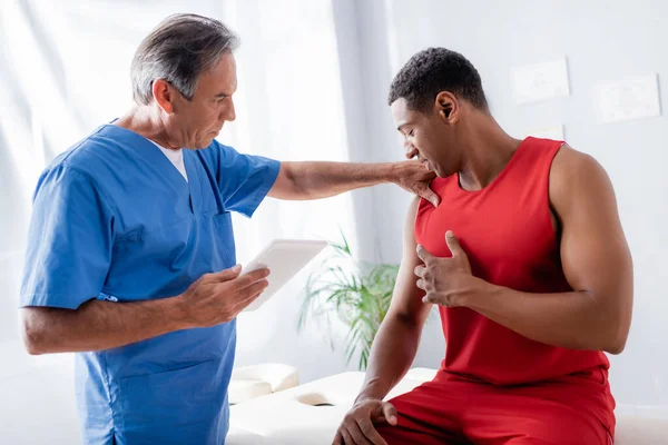 Chiropractor holding digital tablet while working with african american patient in sportswear — Stock Photo