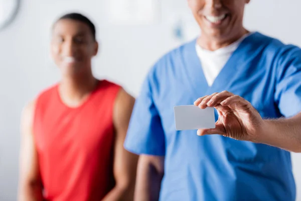 Joyful doctor holding blank card near african american patient in sportswear on blurred background — Stock Photo