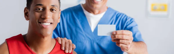 Blurred doctor holding blank card and touching shoulder of happy african american patient, banner — Stock Photo