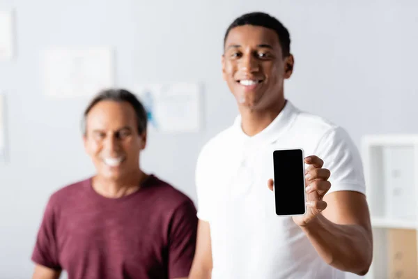 Alegre afroamericano terapeuta celebración de teléfono inteligente con pantalla en blanco cerca de paciente en fondo borroso - foto de stock