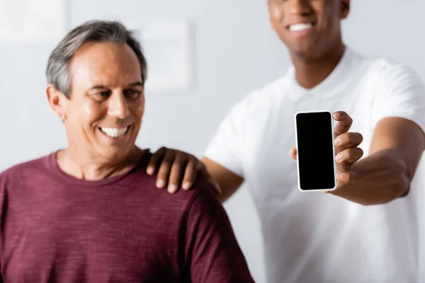 Heureux thérapeute afro-américain tenant smartphone avec écran vide près du patient sur fond flou — Photo de stock