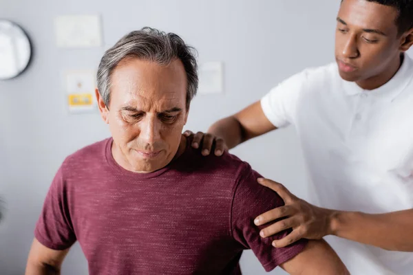 Kinésithérapeute afro-américain flou travaillant avec un homme mûr à la clinique — Photo de stock