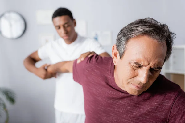 Reifer Mann, der unter Schmerzen leidet, während er von einem amerikanischen Physiotherapeuten auf verschwommenem Hintergrund massiert wird — Stockfoto